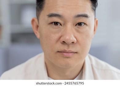 A detailed close-up portrays a middle-aged Asian man with a contemplative, serious expression, captured indoors with a soft focus background. - Powered by Shutterstock