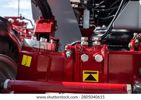 Detailed close-up of a red-painted tractor implement showing metal linkage, hydraulic fittings, mounting bolts, warning decals, and part of a wheel. The image