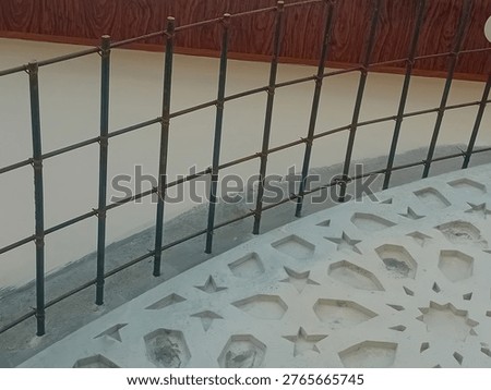 ​This detailed close-up shot captures the complex layering of architectural elements on a building's exterior during the final stages of construction. The central focus is a grid of dark metal rebar frames tied securely with thick wire, which extends from a cream-colored wall, serving as a structural base for decorative cladding. Above the metalwork, a mahogany-patterned PVC ceiling panel provides a rich, warm wood grain aesthetic to the roof eave, featuring an integrated modern recessed downlight. In the foreground, an ornate decorative partition with a geometric star cutout pattern adds a traditional architectural touch to the contemporary facade design. The photograph effectively contrasts the industrial strength of the structural steel with the smooth, finished look of the decorative materials. This visual is ideal for architectural documentation, construction portfolios, and publications focusing on modern exterior building finishes, material integration, and the fusion of traditional patterns in modern design.