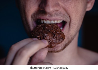 A detailed close-up shot of the mouth of a young handsome man who bites a cookie, holding it in his hand on a home background. Man eating chocolate cookie with peanut, white teeth, close up face. 