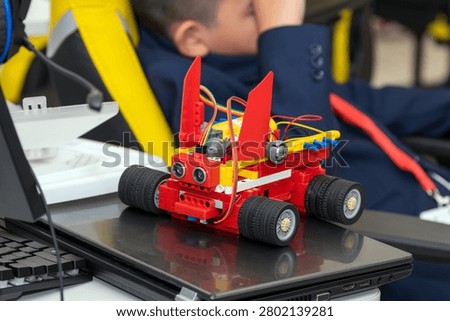 A detailed close-up of a small handmade robotic car with ultrasonic distance sensors and wires standing on a glossy laptop, with a blurred child in a suit in the background. Custom smart robot car with sensors on laptop keyboard