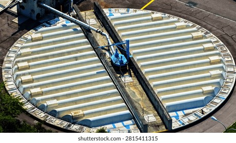 Detailed elevated view of a large covered circular basin at an industrial water or wastewater treatment facility. Repeating curved roof sections create a strong radial pattern across the tank, while a central concrete service bridge, metallic pipeline and blue mechanical equipment reveal the technical structure of the installation.