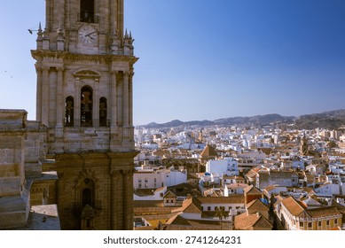 Detailed high angle view featuring the Malaga Cathedral bell tower and tower clock with the urban city skyline and distant mountains under a clear blue sky in Andalusia, southern Spain.