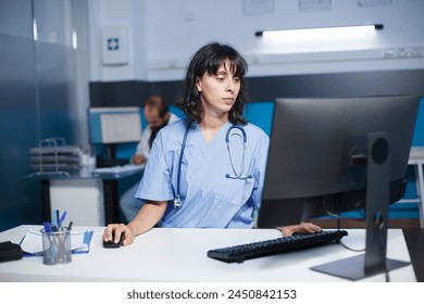 Detailed image of a caucasian nurse operating a desktop computer in a hospital office. A female practitioner wearing blue scrubs checks medical files on a computer in a clinic room. - Powered by Shutterstock