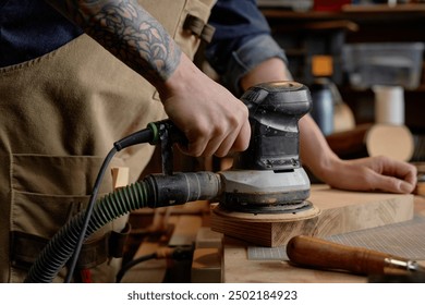 Detailed image showing craftsman using electric sander on wooden piece in workshop. Forearm with tattoo visible, emphasizing intricate work on smoothening wood surface - Powered by Shutterstock