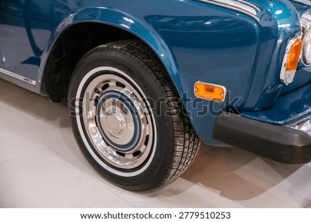 Detailed low-angle view captures front wheel and fender of vintage automobile in glossy blue. Classic chrome hubcap and white stripe tire enhance the vehicle's pristine condition in showroom setting.