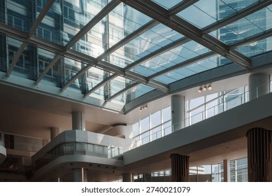 A detailed low-angle view looking through a large glass skylight at a contemporary multi-story office building. The bright interior atrium features white structural columns and reflective glass panels under a clear sky.