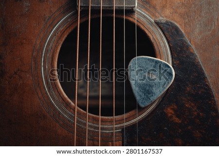 Detailed macro shot of a worn, rustic wooden guitar. A used blue plectrum rests by the soundhole. The warm, dark tones evoke a sense of nostalgia, perfect for folk, blues, or country music themes.