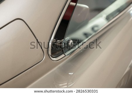 Detailed side view close up shot showing the open rear vent window, chrome window frame trim, hinge mechanism, and fuel door on a vintage champagne metallic coupe bodywork indoors.