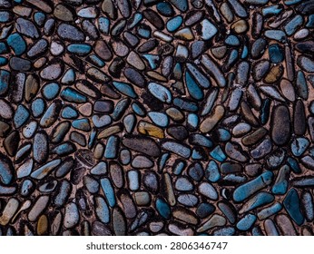 A detailed, top-down full-frame shot capturing the rich and moody texture of an architectural wash pebble floor pavement. The dense surface pattern is formed by countless small, rounded river pebbles and smooth natural stones embedded firmly into a solid concrete matrix. This particular composition features an enhanced cinematic color grading, showcasing a captivating blend of deep teal, rich turquoise, slate grey, and dark earthy tones with strong shadowy contrasts. Each stone displays a unique smooth shape and an organic orientation, creating an excellent non-slip appearance with deep visual depth. This seamless-looking abstract backdrop is highly relevant for modern landscaping designs, luxury pool decks, outdoor patio flooring, tropical garden paths, and high-quality 3D material rendering overlays.