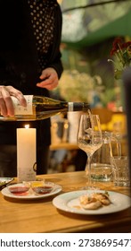 A detailed vertical shot of a server in a patterned shirt and dark cardigan pouring white wine into a glass. The wooden table is artfully arranged with small tasting plates, a lit pillar candle, and a gourmet appetizer. The warm, soft bokeh in the background captures the bustling yet sophisticated atmosphere of a modern restaurant, making it ideal for food and beverage advertising, wine lists, and hospitality industry articles.