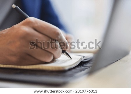 Detailed view of a man's hand jotting down notes in a notepad on a modern laptop, with a blurred background