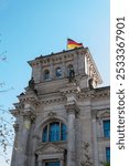 A detailed view of the Reichstag building in Berlin, Germany, with the German flag waving atop the structure against a clear blue sky.