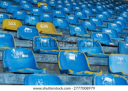 A detailed view of rows of worn out blue and yellow plastic seats in a sports arena or stadium grandstand showing numbers and weathered texture.