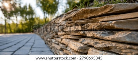 A detailed view of a rustic stone wall bordering a paved walkway, surrounded by lush greenery and trees.