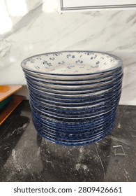 A detailed view of a stack of small, decorative ceramic plates with a distinct blue rim and delicate white-on-blue and blue-on-white floral patterns. The stack is neatly arranged on a dark, reflective marble counter, with a white marble wall providing the background. This high-quality commercial stock photo is suitable for homeware, retail, dining, and decor publications and marketing.