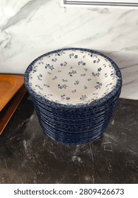 A detailed view of a stack of small, decorative ceramic plates with a distinct blue rim and delicate white-on-blue and blue-on-white floral patterns. The stack is neatly arranged on a dark, reflective marble counter, with a white marble wall providing the background. This high-quality commercial stock photo is suitable for homeware, retail, dining, and decor publications and marketing.