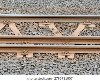 Detailed view of two parallel rust-colored steel rails mounted on concrete sleepers with metal clips and fastenings, surrounded by gray crushed stone ballast