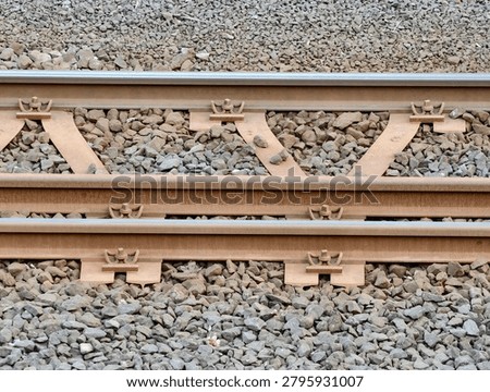 Detailed view of two parallel rust-colored steel rails mounted on concrete sleepers with metal clips and fastenings, surrounded by gray crushed stone ballast