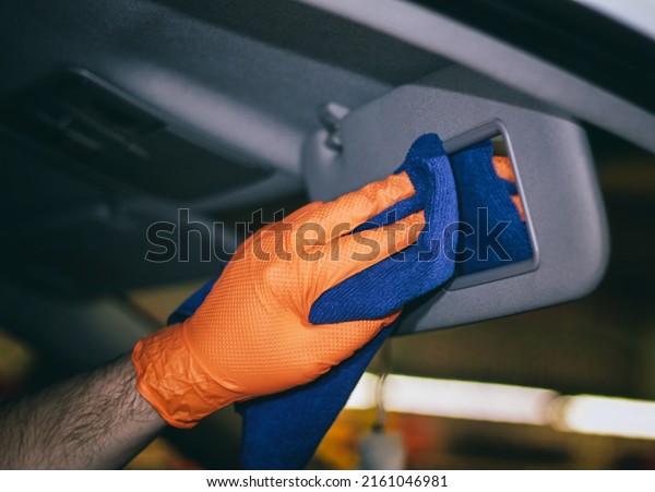Detailing and car\
cleaning services, the concept of car washing and cleaning. A male\
worker in orange rubber gloves is cleaning the car sun visor mirror\
with a blue microfiber\
towel.	