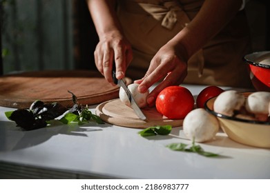 Details: Chef's hands using a kitchen knife, slicing fresh mushroom champignon on a chopping board. Fragrant basil leaves lying on a kitchen countertop in a rustic kitchen . Chopped pizza ingredients.