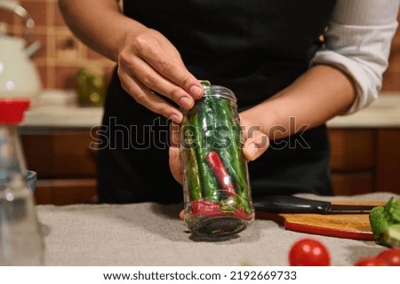 Details: Hands of a housewife in black chef's apron holding a sterilized jar filled with fresh organic red and green spicy chili peppers, prepared for marinating. Canning and preserving food concept
