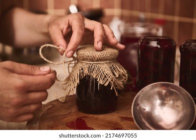 Details: Hands of a housewife, woman chef confectioner tying bow with a rope around a burlap on a lid of a jar with freshly made cherry berry jam at home kitchen. Organic homemade preserved product