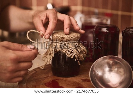 Details: Hands of a housewife, woman chef confectioner tying bow with a rope around a burlap on a lid of a jar with freshly made cherry berry jam at home kitchen. Organic homemade preserved product