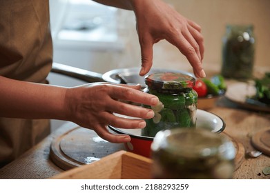 Details: housewife's hands cover with a sterilized lid a glass jar with freshly pickled organic cucumbers. Canning, marinating, pickling concept. Preparing homemade marinated delicacies for the winter