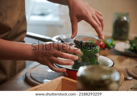 Details: housewife's hands cover with a sterilized lid a glass jar with freshly pickled organic cucumbers. Canning, marinating, pickling concept. Preparing homemade marinated delicacies for the winter