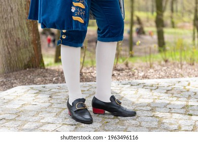 Details of a man's dress in a baroque costume. White stockings and black shoes, golden buttons, decorative hems. A man standing on a cobbled walkway in the background of a blurred garden backdrop.