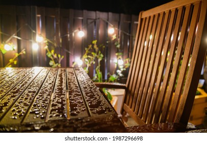 Details Of Some Water Drops On A Table And Some Wood Chairs After A Summer Rain In The Backyard Garden In The Middle Of The Night With Light Garland Bulbs In Background. Design Ideas.