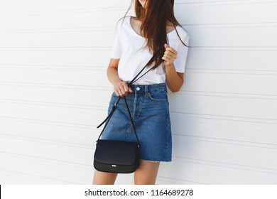 Details of trendy casual summer or spring outfit. Woman wearing blue denim mini skirt, white tshirt, small black cross body bag standing near white roller door. Everyday look. Street fashion. No face.