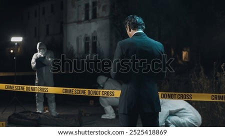 Detective inspecting area and taking notes in notebook at crime scene at night as forensic experts collect evidence behind police tape next to headless body