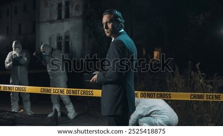 Detective inspecting area and taking notes in notebook at crime scene at night as forensic experts collect evidence behind police tape next to headless body