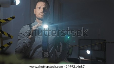 A detective man with a beard inspects a dark indoor crime scene using a flashlight.