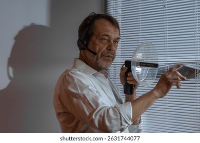 Detective on assignment. A man in a headset stands at a window with blinds, conducting covert surveillance