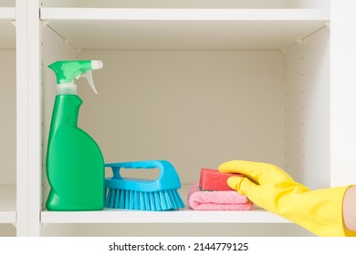 Detergent bottle, brush, rags and sponge on white shelves inside opened wardrobe. Woman hand in rubber protective glove taking cleaning things. Closeup. Front view.