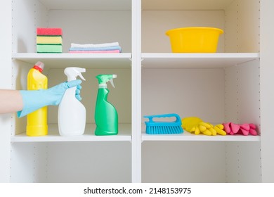 Detergent bottles, bowl, brush, rags and sponges on white shelves inside opened wardrobe. Woman hand in rubber protective glove taking cleaning things. Closeup. Front view.
