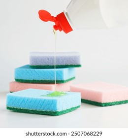 Detergent from a white plastic bottle with a red cap is poured onto multi-colored sponges for washing dishes and cleaning. White background. Housekeeping and cleaning. Photo. Copy space. Close-up - Powered by Shutterstock