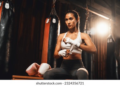 Determined Asian female Muay Thai boxer with muscularity physical readiness body wraps her hand and dons or wearing boxing glove, preparing for intense boxing training in the ring at gym. Impetus - Powered by Shutterstock