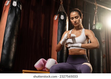 Determined Asian female Muay Thai boxer with muscularity physical readiness body wraps her hand and dons or wearing boxing glove, preparing for intense boxing training in the ring at gym. Impetus - Powered by Shutterstock