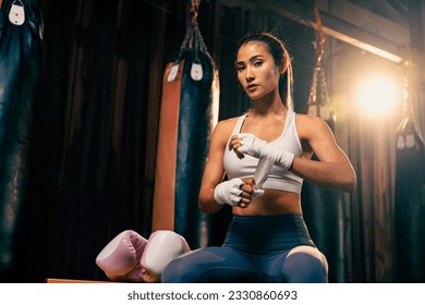 Determined Asian female Muay Thai boxer with muscularity physical readiness body wraps her hand and dons or wearing boxing glove, preparing for intense boxing training in the ring at gym. Impetus - Powered by Shutterstock