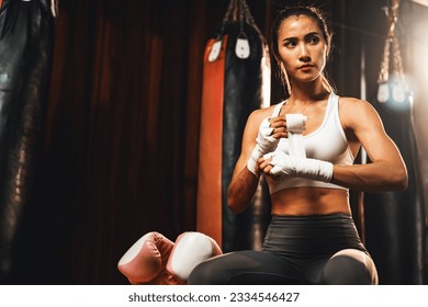 Determined Asian female Muay Thai boxer with muscularity physical readiness body wraps her hand and dons or wearing boxing glove, preparing for intense boxing training in the ring at gym. Impetus - Powered by Shutterstock