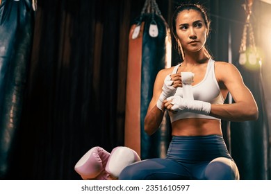 Determined Asian female Muay Thai boxer with muscularity physical readiness body wraps her hand and dons or wearing boxing glove, preparing for intense boxing training in the ring at gym. Impetus - Powered by Shutterstock