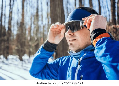 Determined athlete in a snowy forest, putting on stylish sunglasses before training in the winter sun. Blue jacket and orange headband stand out against white snow, showing commitment to fitness - Powered by Shutterstock