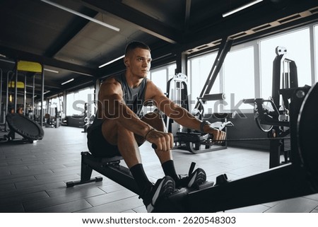 Determined attractive male using rowing machine in contemporary gym. Fit young man working out on rowing equipment at fitness center. Weight loss and cardio exercise concept