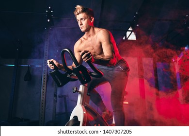 Determined bare-chested sportsman engaged in a bicycle simulator in the gym, warming up before powerlifting workout