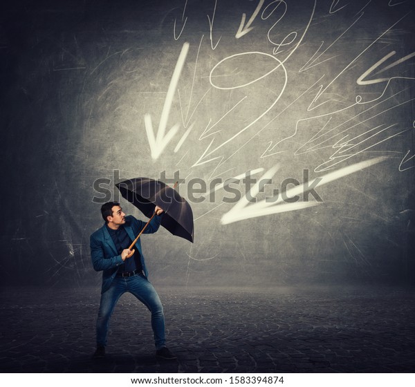Determined businessman hiding behind umbrella\
as a shield to protect of any danger and high pressure of arrow\
sketches pointed to him. Business concept of facing adversity and\
auto defending.