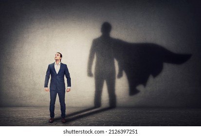 Determined businessman stands confident in a hero stance and casting a brave superhero shadow on the wall behind. Business leadership and motivation concept. Ambition and strength symbols
